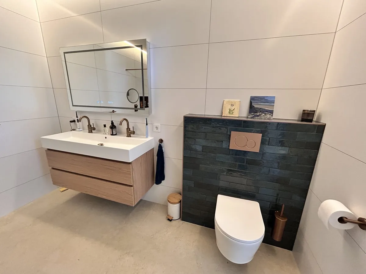 Hangend toilet met een koperen bedieningsplaat voor een groene tegelwand, naast een badkamermeubel