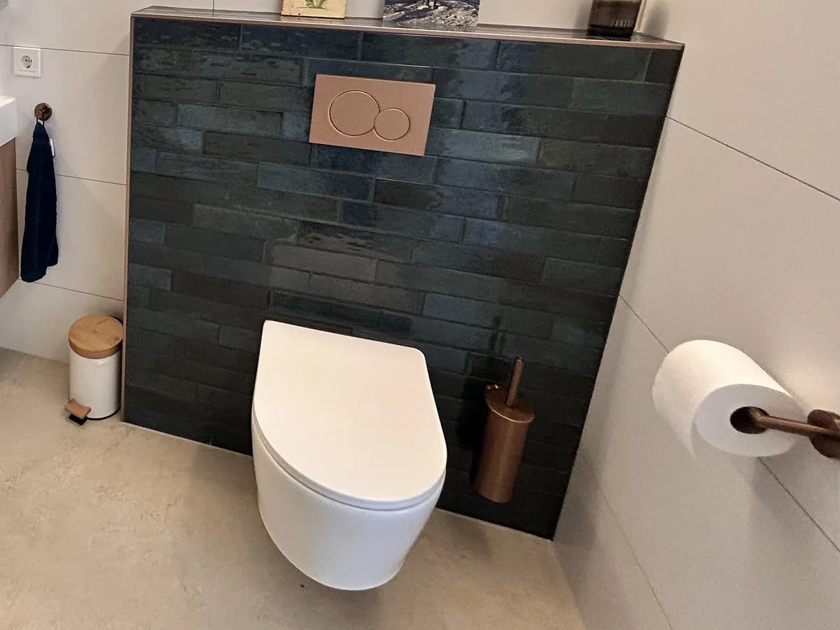 Hangend toilet voor een wand met donkergroene tegels en een koperen bedieningsplaat